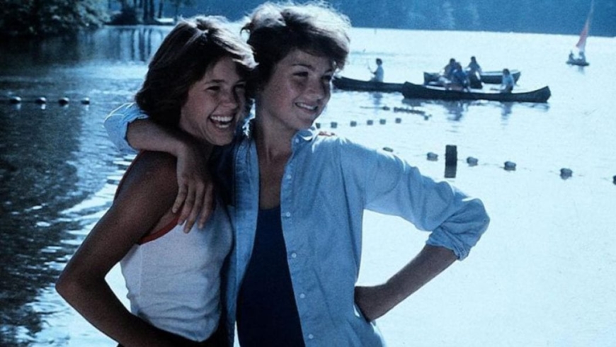 Two women in casual attire sharing a joyful moment by a body of water, with one embracing the other from the side and both smiling brightly, suggesting a close and happy relationship amidst a serene setting with boaters in the background.