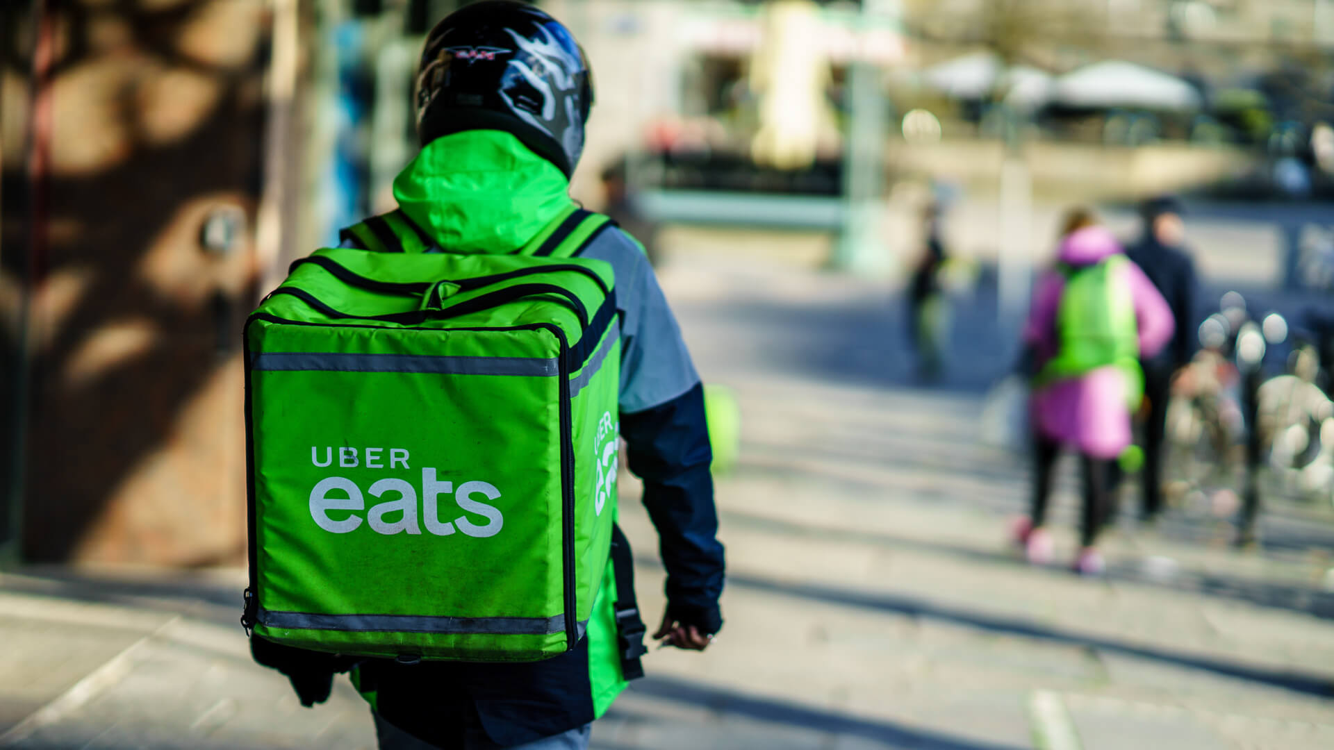 A person walking down a sidewalk with a green Uber Eats backpack.