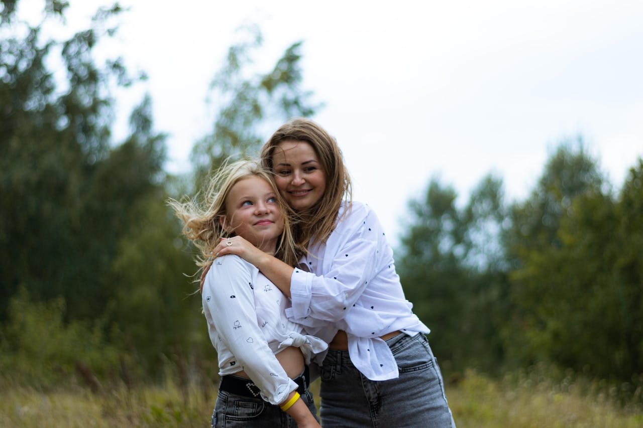 Smiling mother and daughter close together in a grassy field in similar white long sleeve dress and jeans