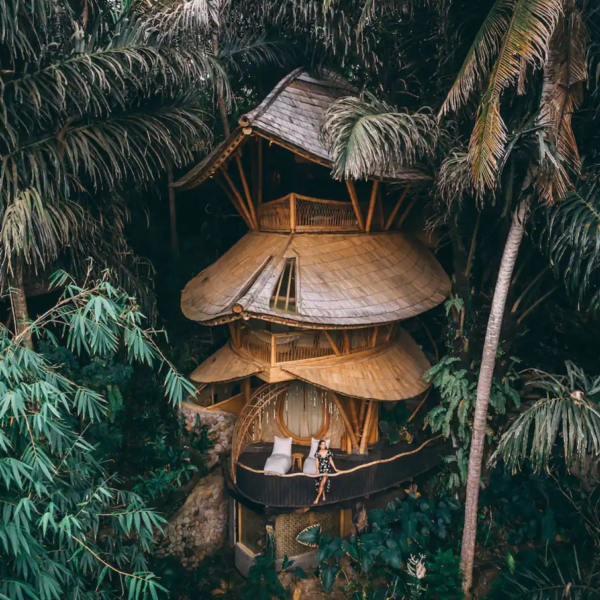 Aerial view of a four-storey tree house nestled among the trees in Bali, with a woman on the balcony