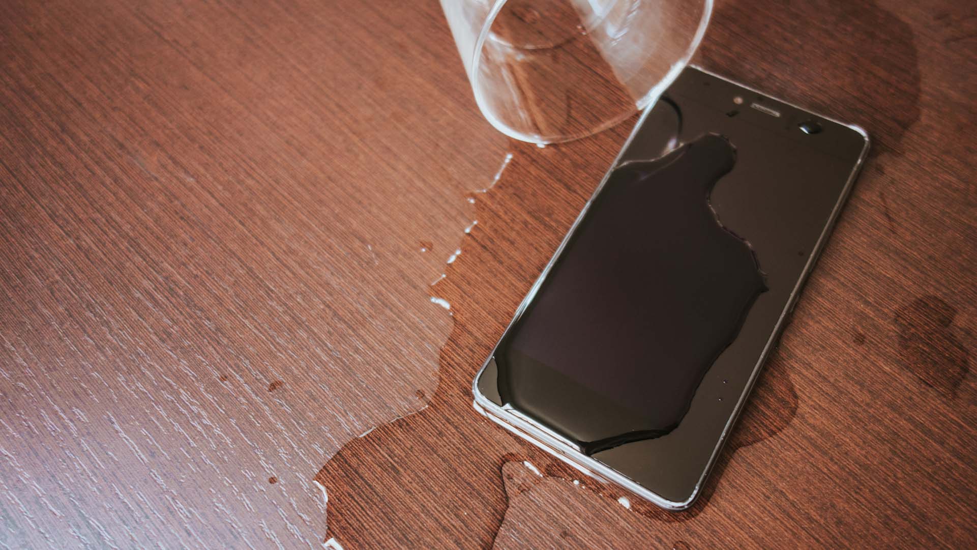 A smartphone lies on a wooden surface with spilled liquid spreading from an overturned glass.