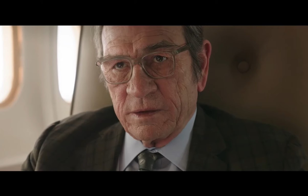 A bespectacled Tommy Lee Jones in a suit and tie aboard a private jet in a scene from ‘The Burial’