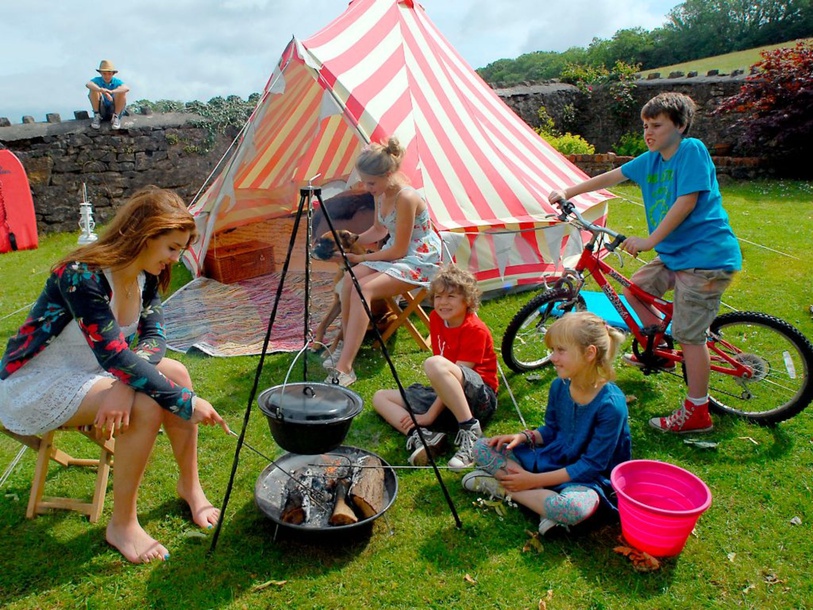 Lively outdoor scene with children engaging in various activities such as cooking over a campfire, biking, and relaxing, suggesting a sense of adventure and family-friendly fun.