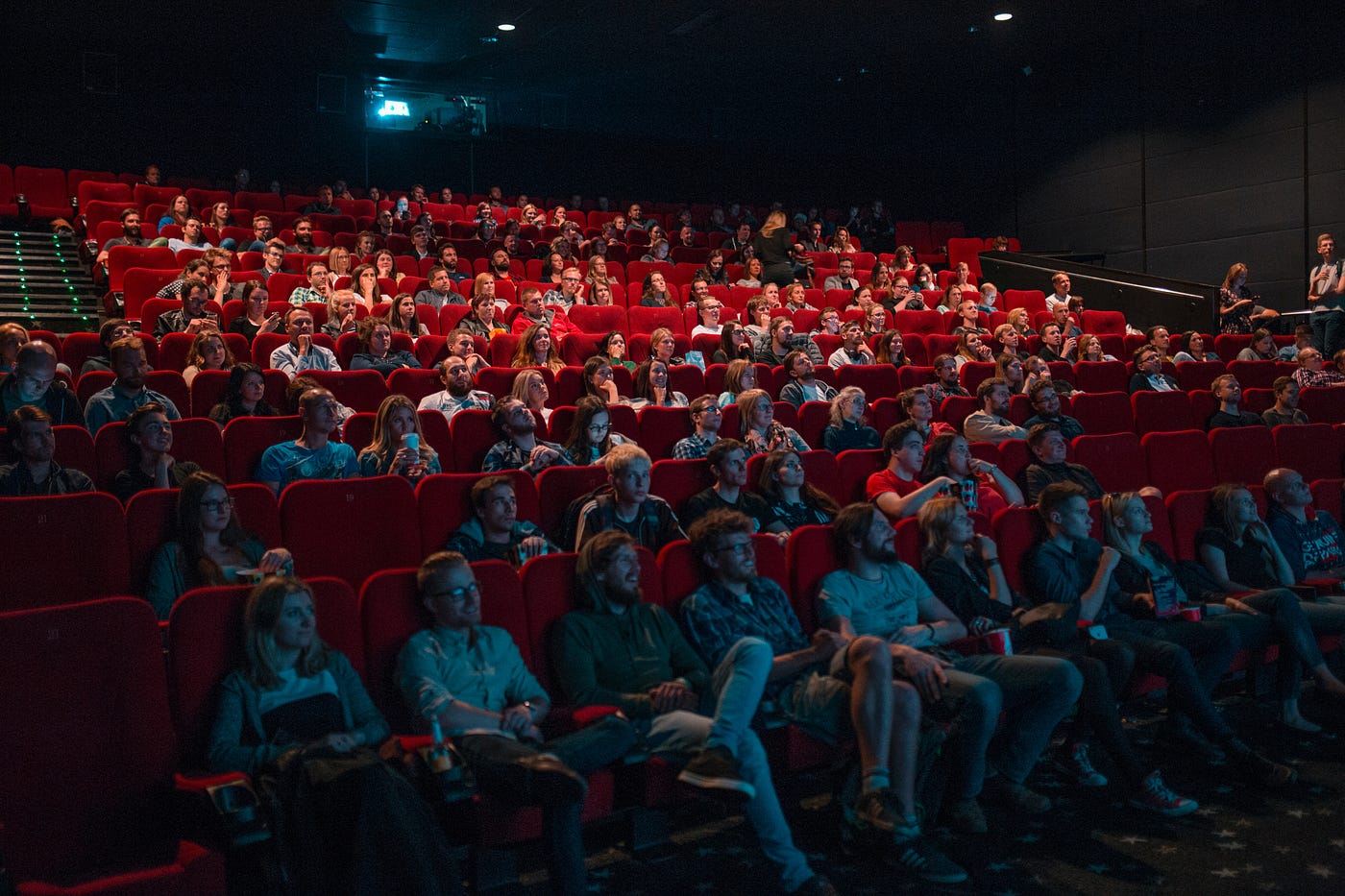 A captivated audience seated in a dimly lit theater with red chairs, watching intently.