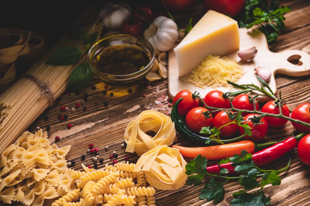 Enticing selection of fresh Italian cooking ingredients, including various pasta shapes, ripe tomatoes, chili peppers, garlic, herbs, a wedge of cheese, and olive oil, all arranged on a rustic wooden surface, evoking the essence of authentic Italian cuisine.