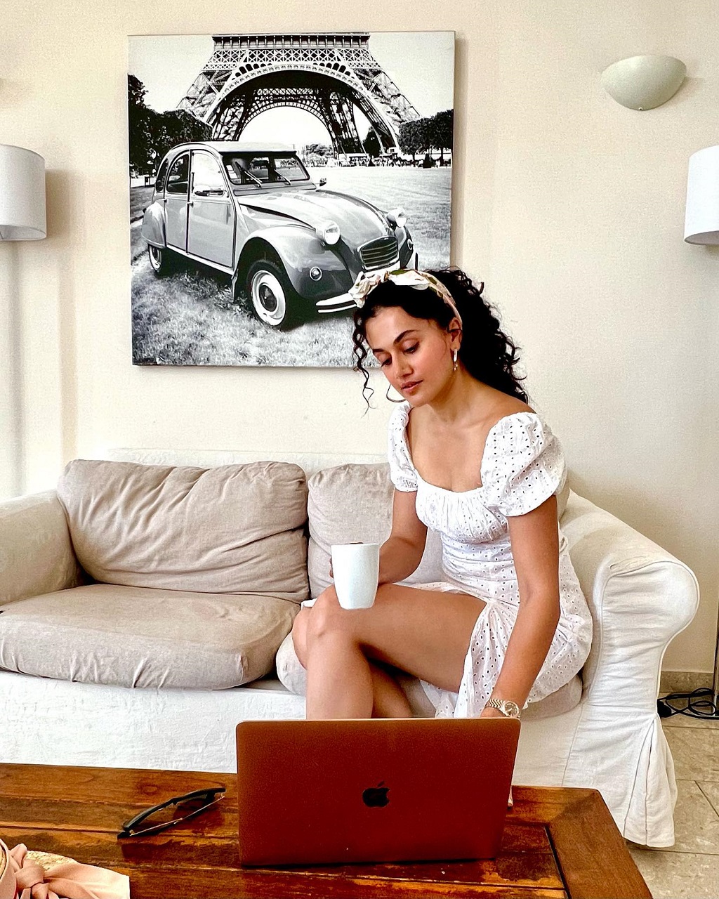 Taapsee Pannu in a white dress seating on a couch while holding a white mug and looking at her MacBook