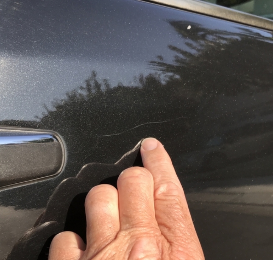 Hand pointing at clear coat scratch of a black car