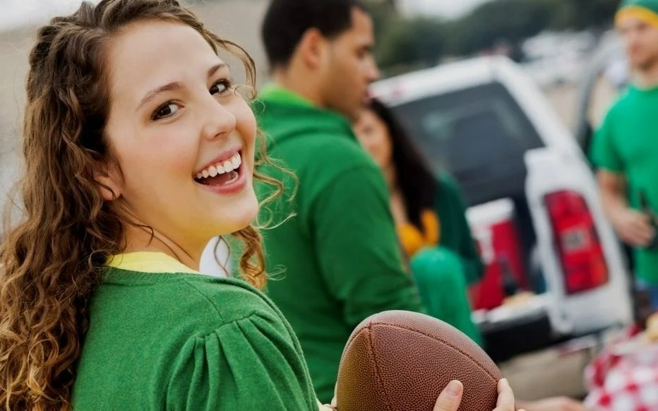 A smiling woman holding a football with friends in a tailgate setting.
