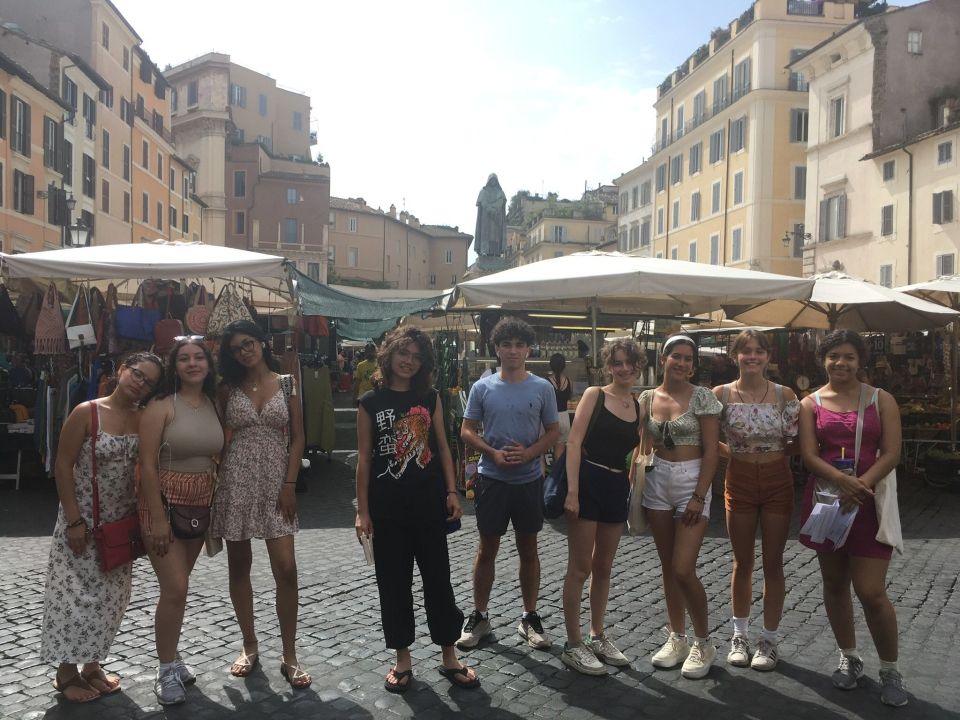 A group of happy young individuals posing for a photo in what appears to be a lively outdoor market square, surrounded by historic buildings and market stalls.