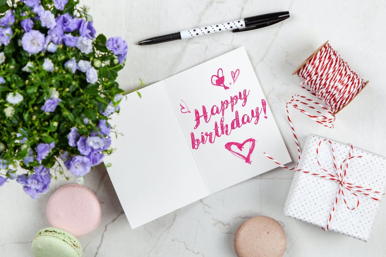 A handwritten ‘happy birthday’ on a white card surrounded by a pen, flower pot, thread, macaroons and gift box