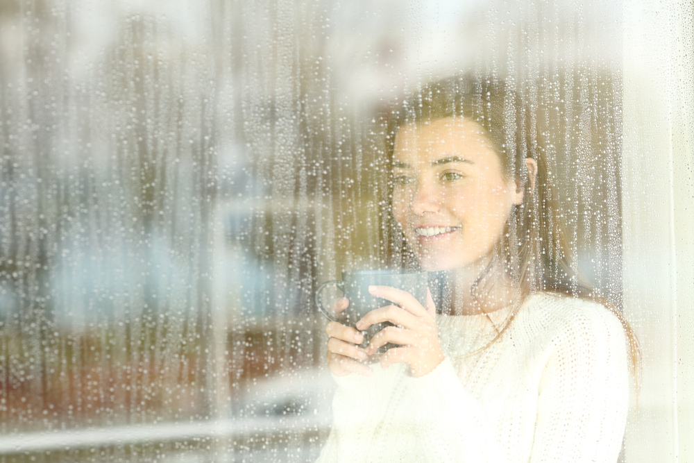 Happy teen watching the rain through a window drinking coffee at home