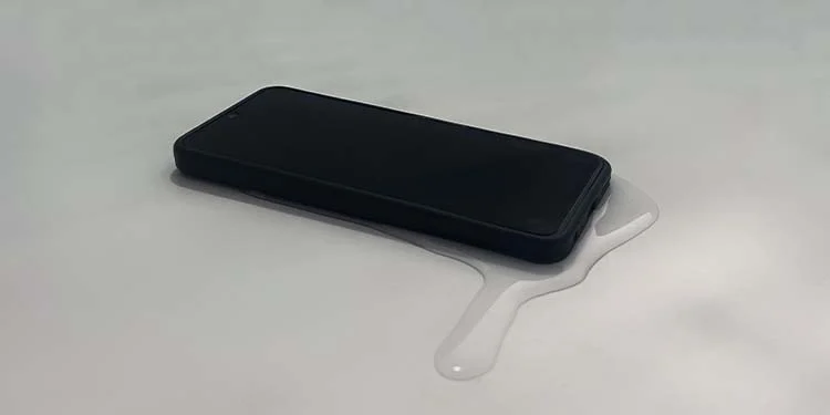 A black smartphone rests on a white surface with a spill of liquid pooling around it.