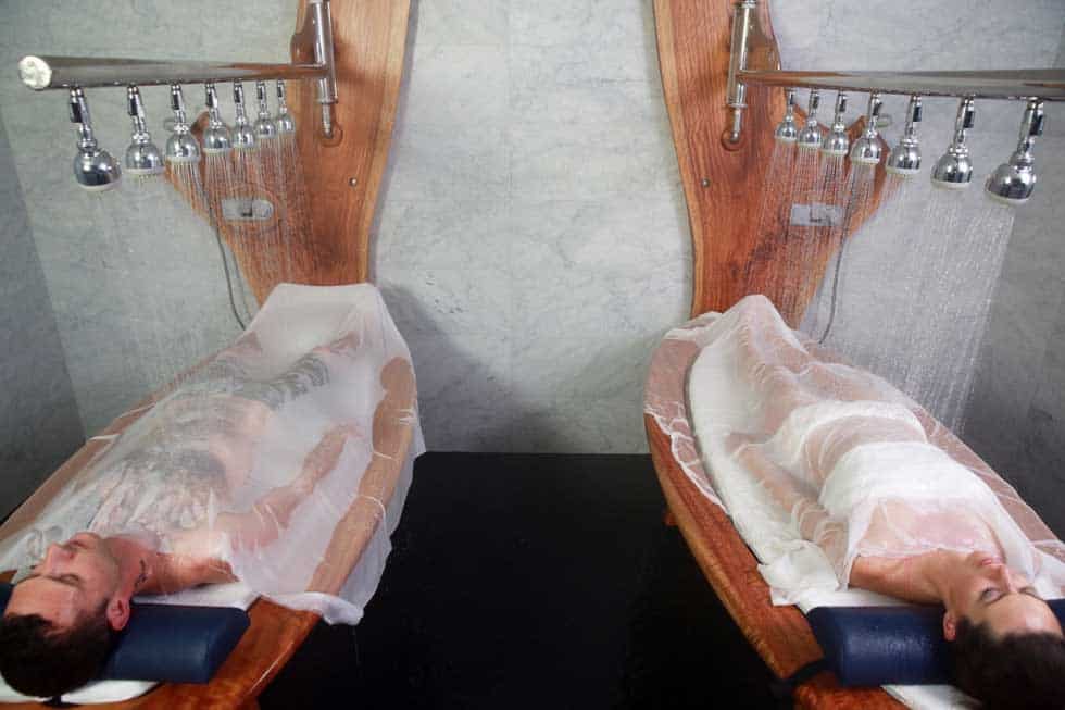 Two individuals lying on massage tables while being showered with multiple streams of water, a treatment often found in high-end spas or therapeutic centers.