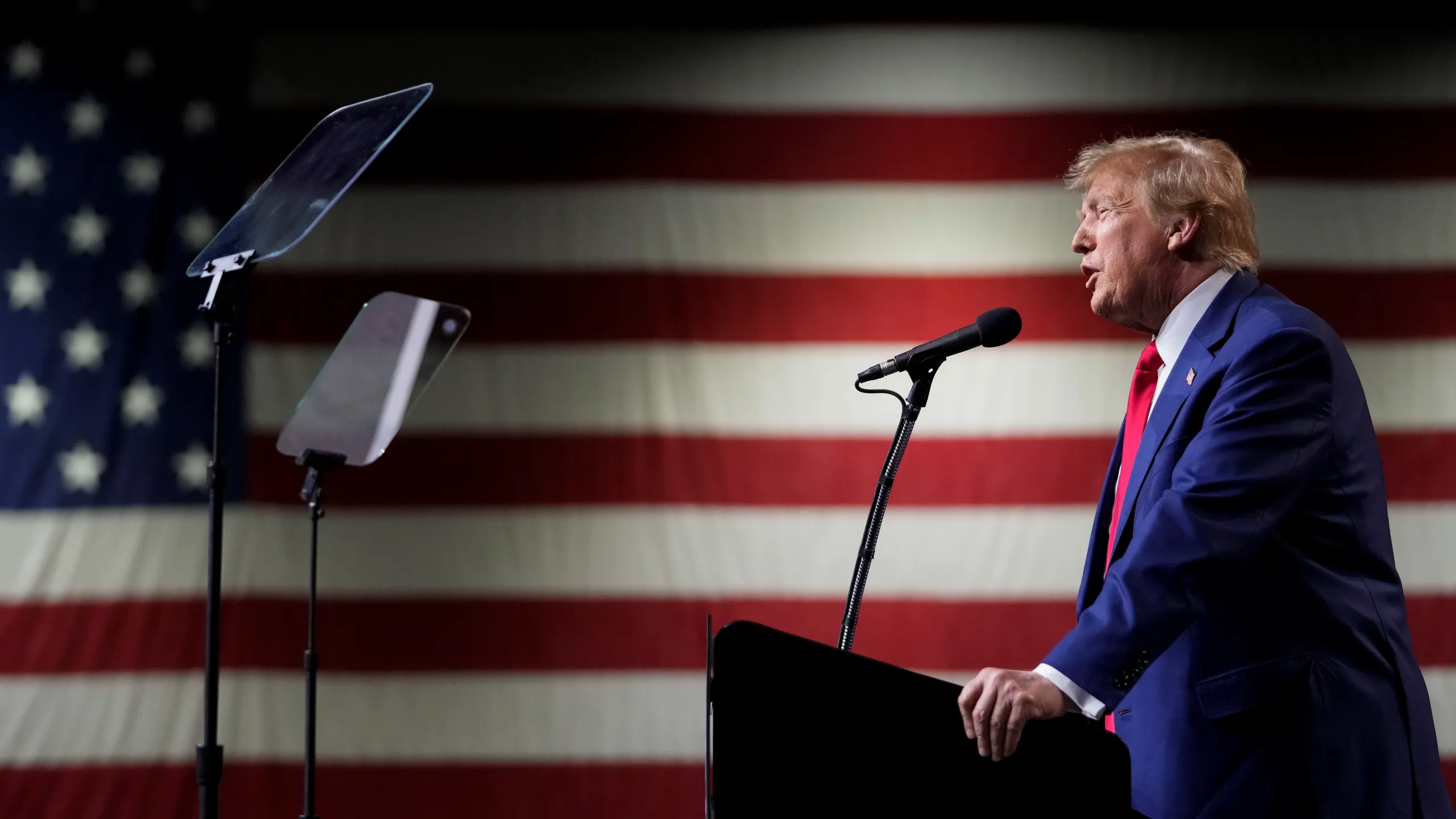 Donald Trump speaking while standing on dice with American flag in background.