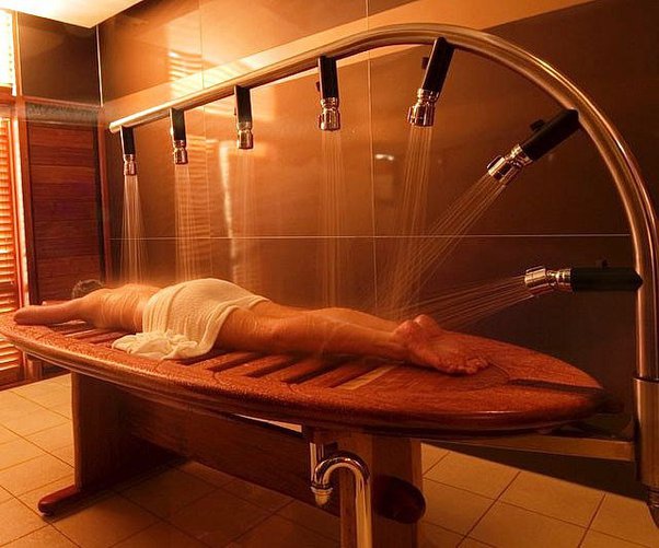 A person lies on a curved wooden table undergoing a multi-shower spa treatment in a dimly lit room.