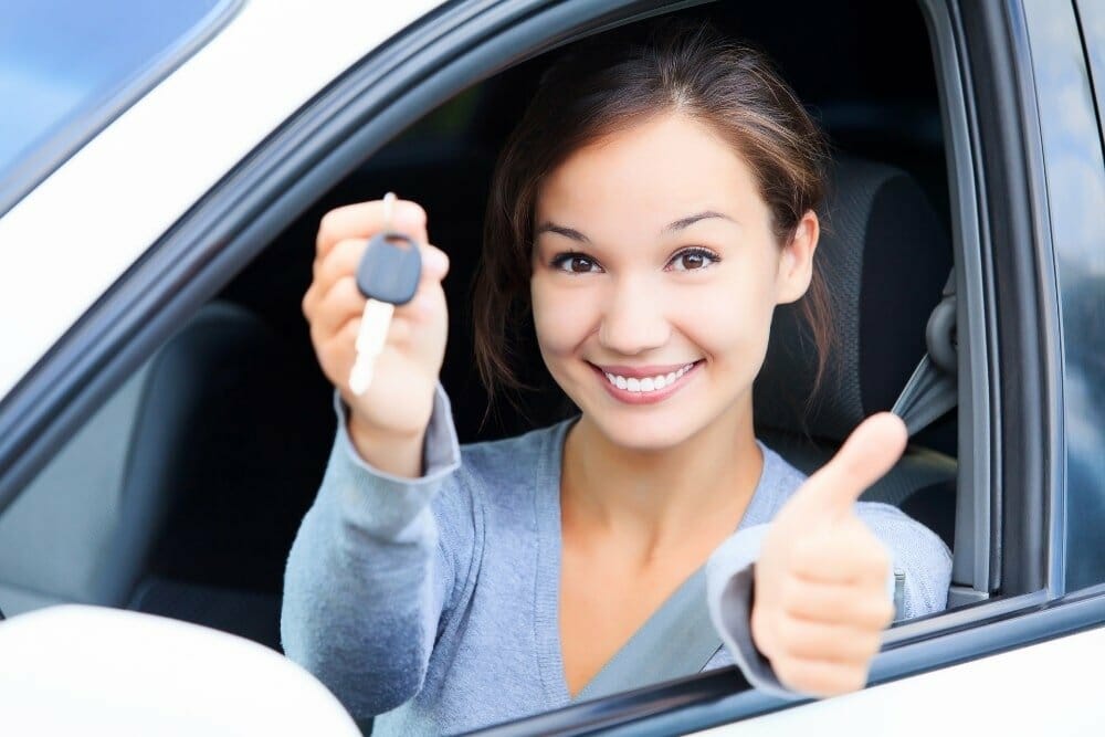 A girl holding car keys while sitting in car giving a thumbs up