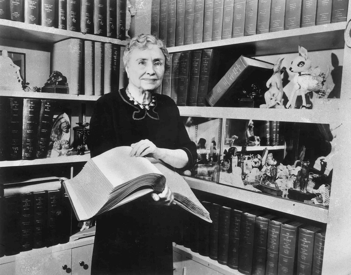 An older woman stands confidently in front of a bookshelf, holding an open book, surrounded by various artifacts and trinkets.