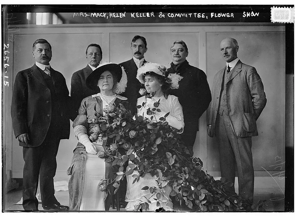 A vintage black-and-white photograph depicts Helen Keller, surrounded by a committee and holding a bouquet, at a flower show event.