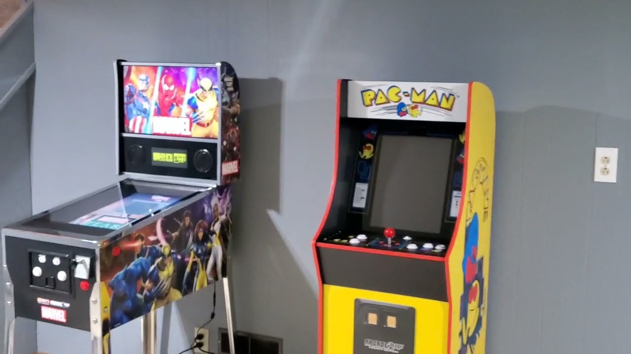 Two arcade machines, one branded with PAC-MAN and the other showcasing colorful characters, stand side by side against a gray wall.