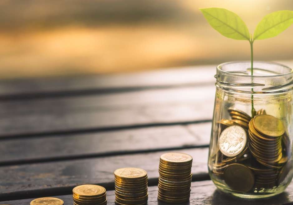A plant growing out of a jar full of coins with additional coins stacked next to it, symbolizing the concept of financial growth or investment.