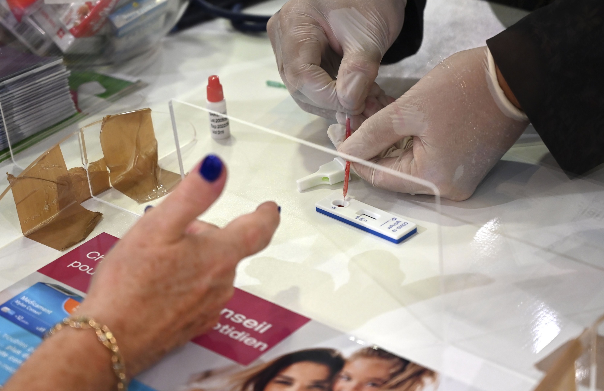 A medical professional conducts a test using a dropper and test strip while a person with a painted fingernail observes.