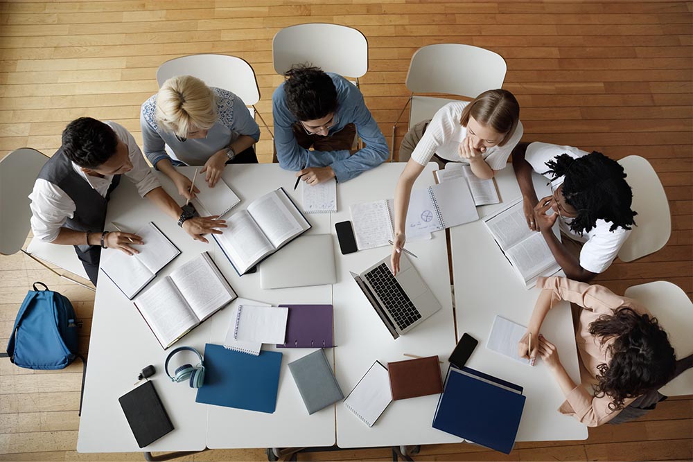 Top-down view of a diverse group of individuals engaged in a collaborative study session around a table with books, notebooks, and laptops.