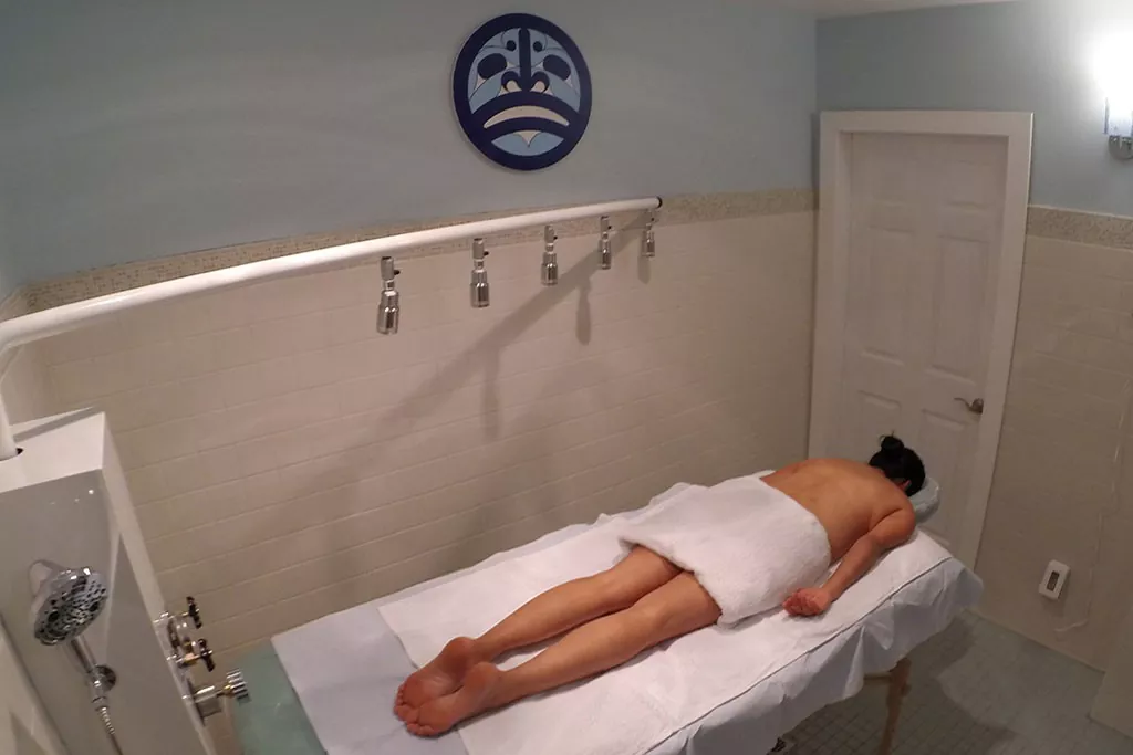 A person lying face down on a massage table in a blue-themed room with a wall-mounted symbol and white fixtures.