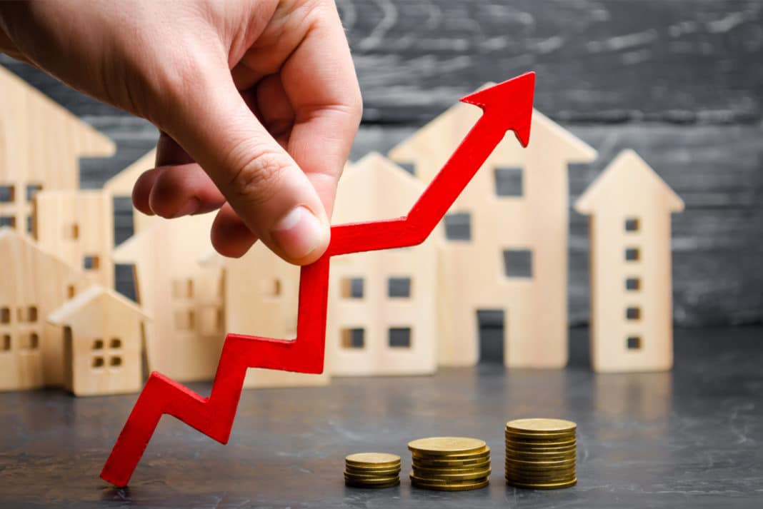 Upward-trending arrow over wooden house models and coins, symbolizing increasing real estate values or investment returns.