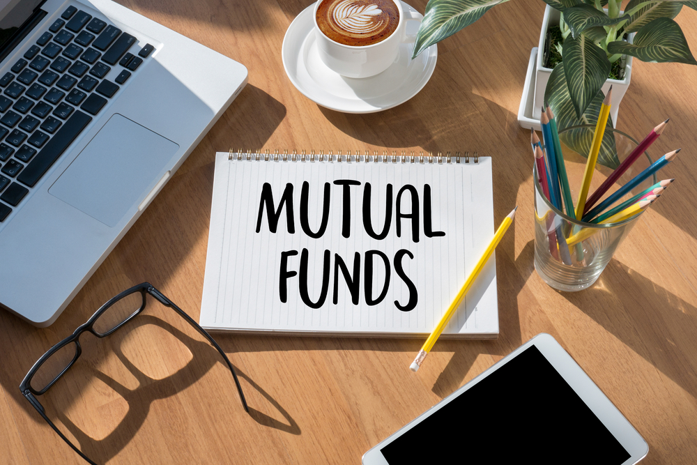 The words mutual funds written on a notebook sitting beside a laptop, tablet, an eyeglss, and a cup filled with pencils