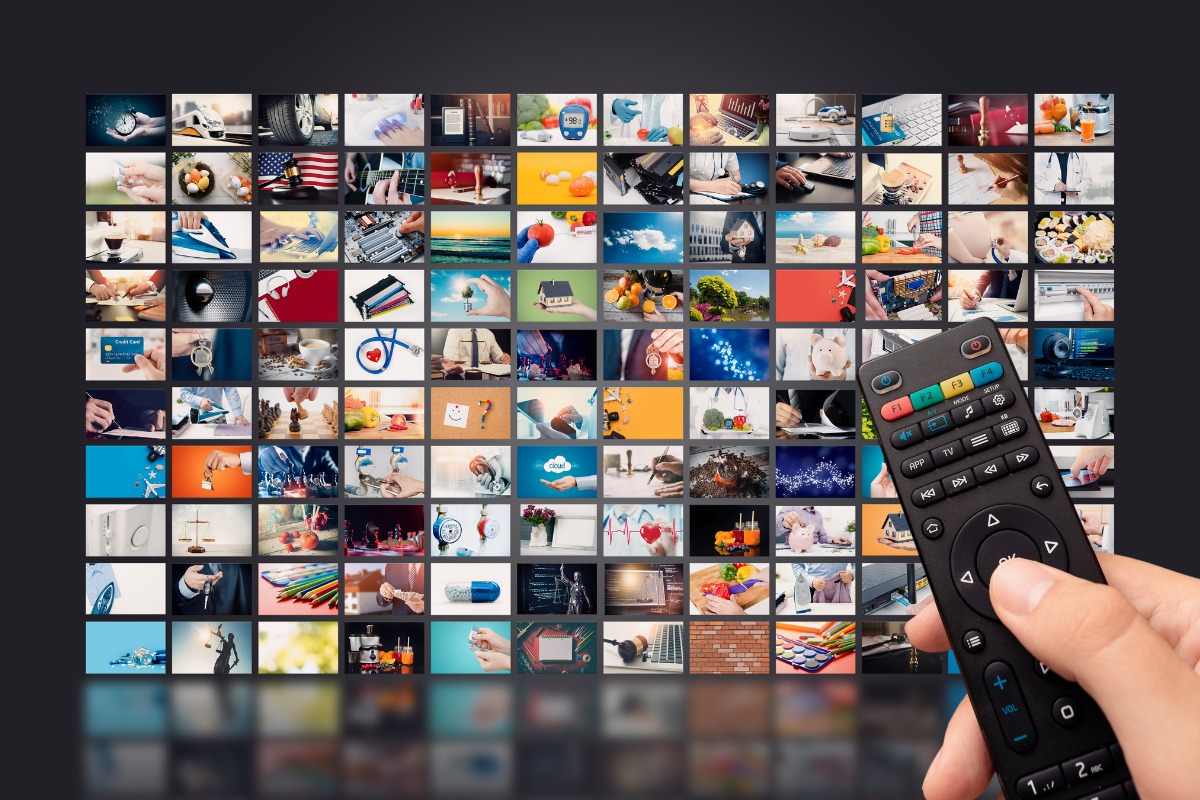 A hand holds a remote control in front of a screen displaying a mosaic of diverse television channels and programs.