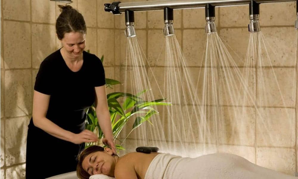 A therapist attending to a relaxed client under multiple showerheads in a tiled spa setting with a plant.