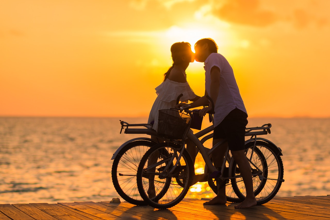  Man Wearing White T-shirt Kissing a Woman While Holding Bicycle on River Dock during Sunset