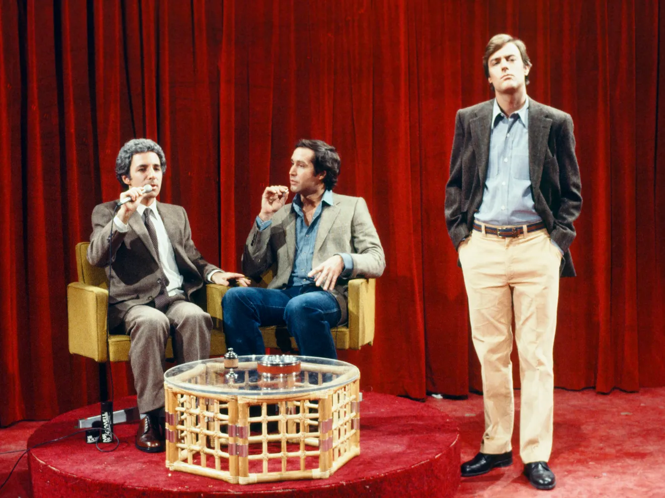 Peter Aykroyd is standing in "Saturday Night Live." while two co cast are sitting on stage.