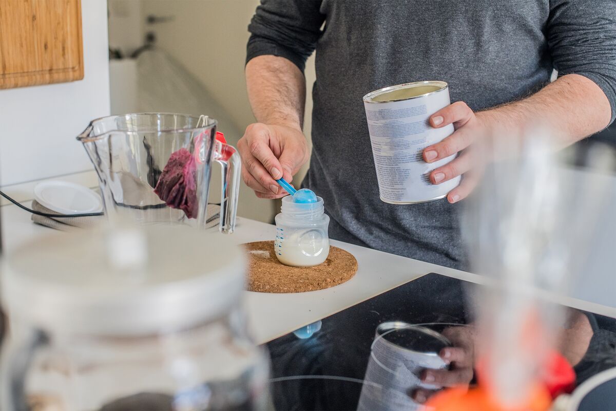 Ndividual is preparing baby formula by scooping powder into a bottle on a kitchen countertop, surrounded by various kitchen items.