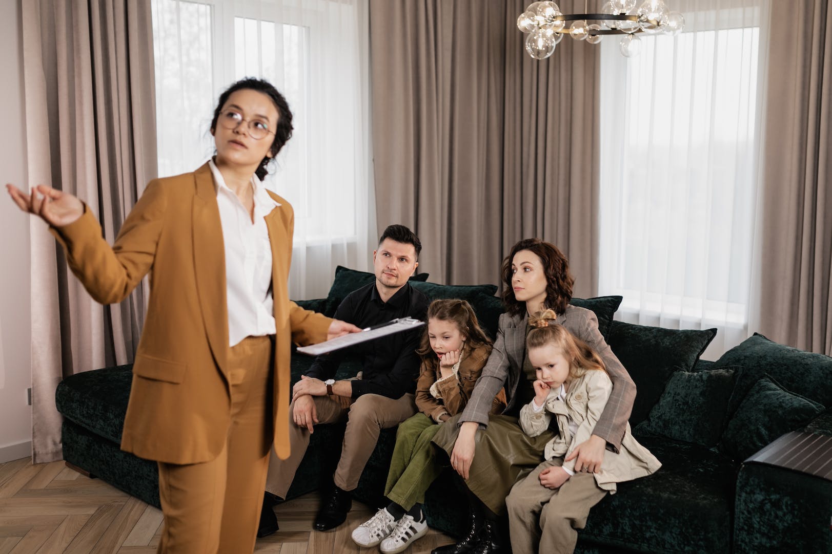 A woman is standing and telling about house to a couple and their two kid who are sitting in couch.