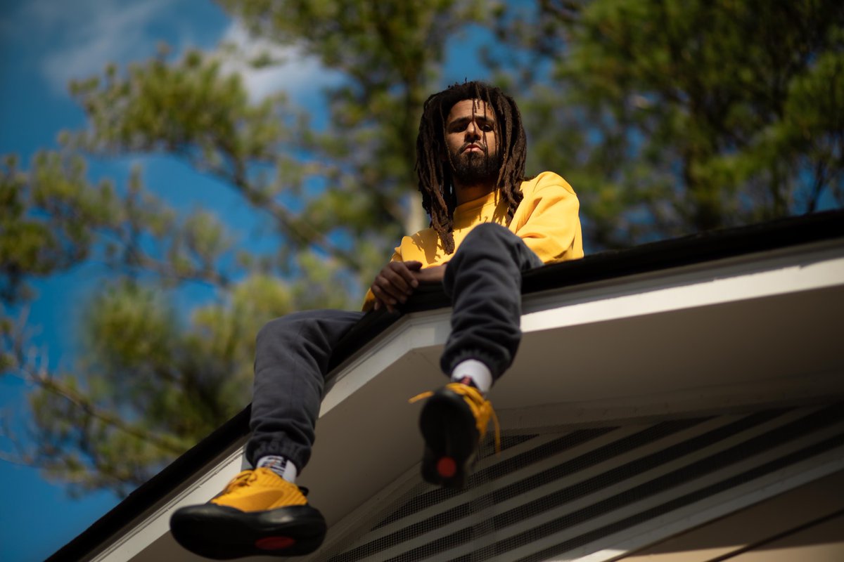 J. Cole wearing a yellow sweater while sitting on a roof
