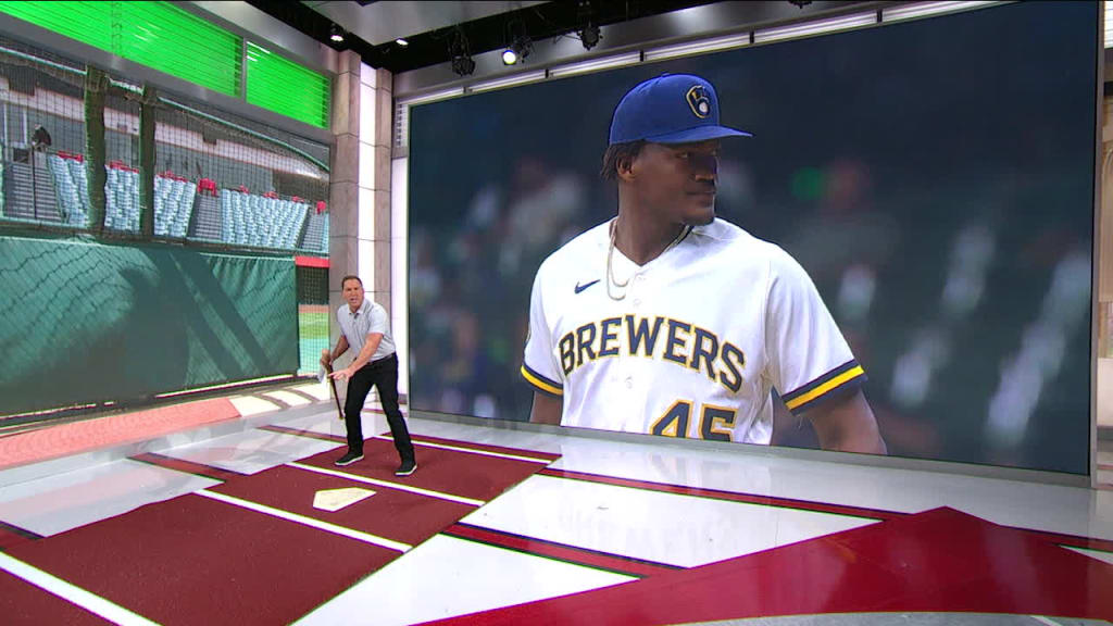 Man standing on a virtual baseball diamond in a studio setting, with a large screen behind him displaying a life-size figure of a player in a Milwaukee Brewers jersey.