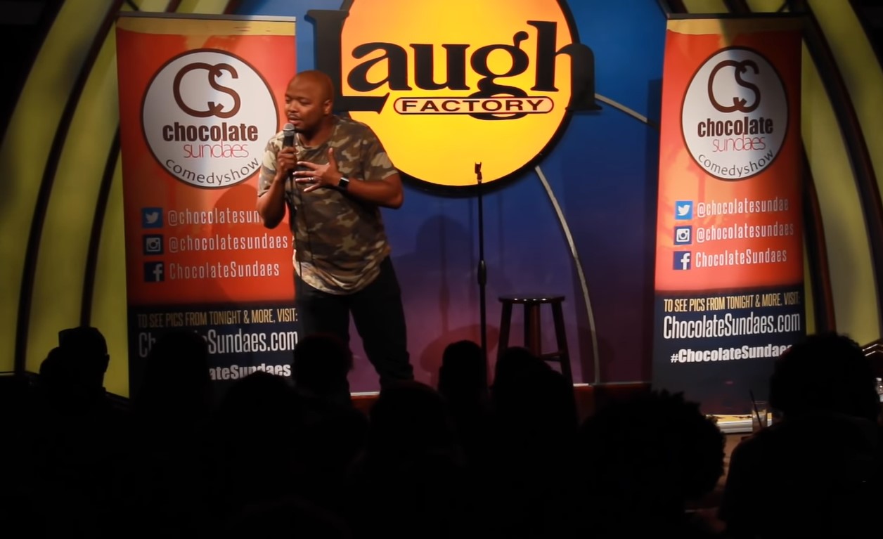 Kev On Stage in camouflage T-Shirt, with left hand on his chest performing near the audience at Laugh Factory