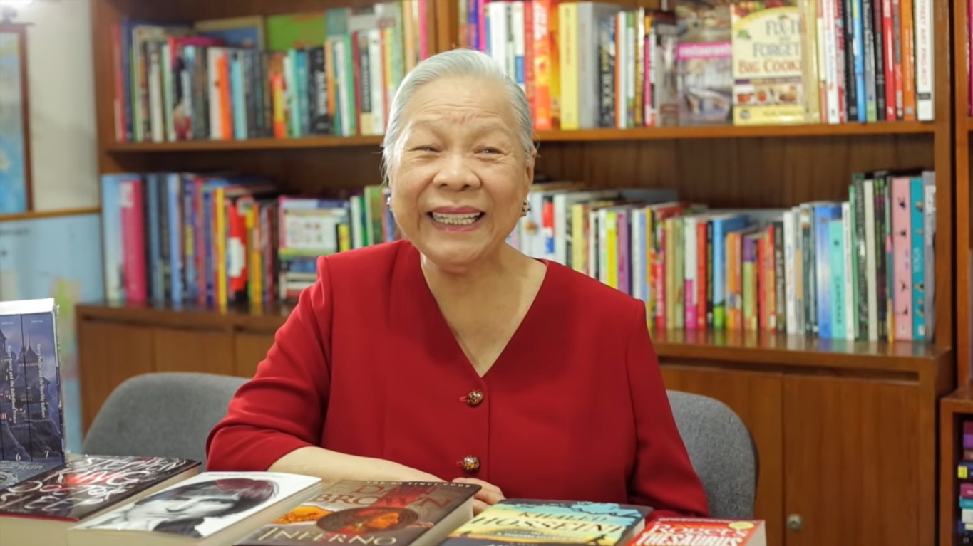 Socorro Ramos in red long sleeve buttoned clothes smiling widely and a bookshelf behind her