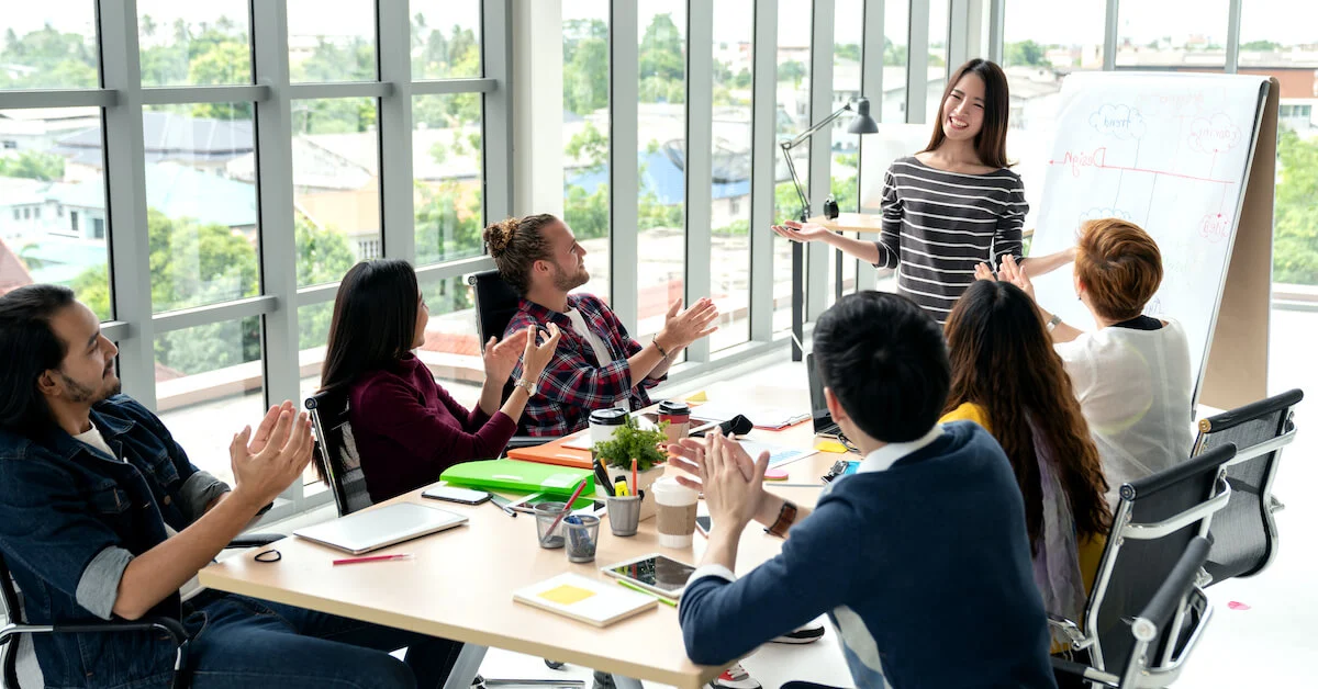 A diverse group of professionals engaged in a meeting, with one person presenting at the whiteboard, highlighting a collaborative and inclusive workplace environment.