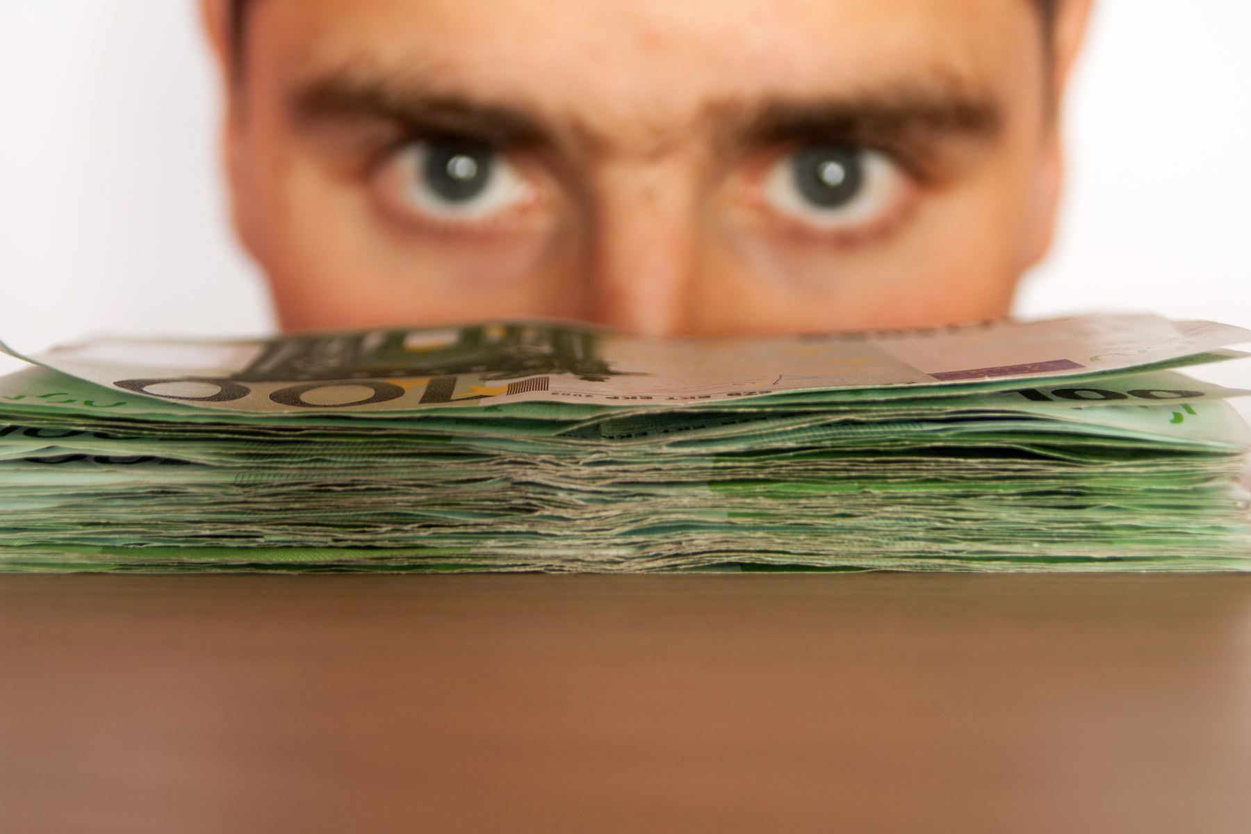 A close-up of a person's eyes peering over a large stack of hundred-dollar bills, conveying a sense of curiosity, scrutiny, or possibly greed.