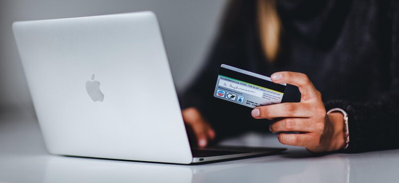 A person holding a credit card in one hand while using a MacBook with the other, a scenario typically associated with online shopping or making an online transaction.