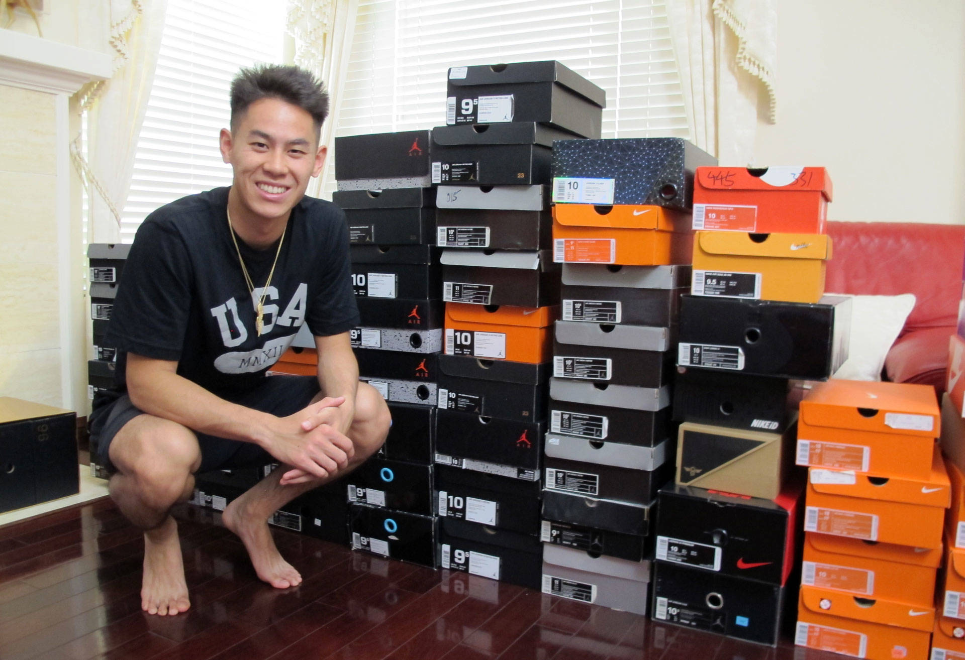A smiling individual sitting next to a large stack of shoeboxes from various popular brands, indicating a collection of footwear.