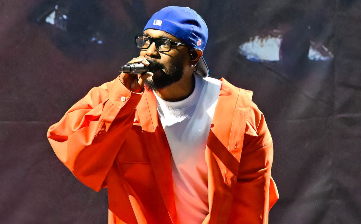 Kendrick Lamar wearing an orange jacket and blue cap while holding a mic