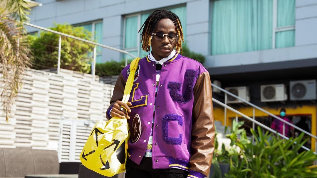 Fireboy DML wearing a purple jacket and glasses while holding a yellow bag